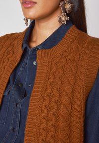 Gilet en tricot marron avec un design en cable, col côtelé, porté sur une chemise en denim bleu avec des boutons noirs. De grandes boucles d'oreilles florales complètent le look.
