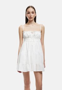 Vestido de verano de algodón blanco con tirantes finos, cuerpo fruncido y falda de capas. Presenta un escote con volantes y un lazo ajustable en la parte delantera.