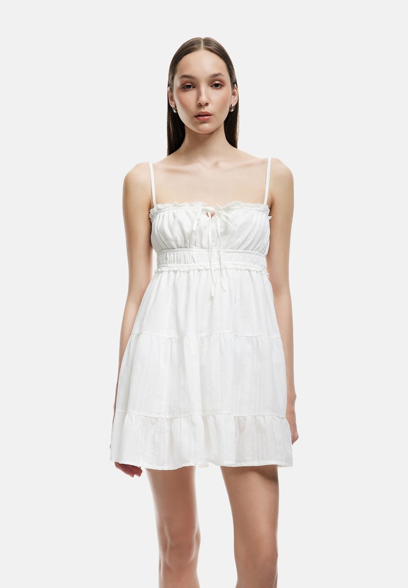 Vestido de verano de algodón blanco con tirantes finos, cuerpo fruncido y falda de capas. Presenta un escote con volantes y un lazo ajustable en la parte delantera.