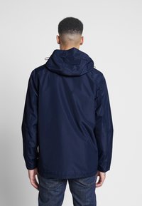 Granatowa wodoodporna kurtka z kapturem, o gładkiej fakturze i regulowanych troczkach, zestawiona z ciemnymi dżinsami. Widok od tyłu.