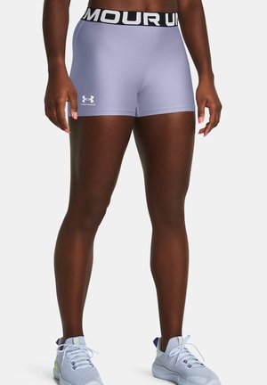 Personne en forme portant un short de compression Under Armour violet clair avec une ceinture noire et des chaussures de course gris clair, debout sur un fond blanc.