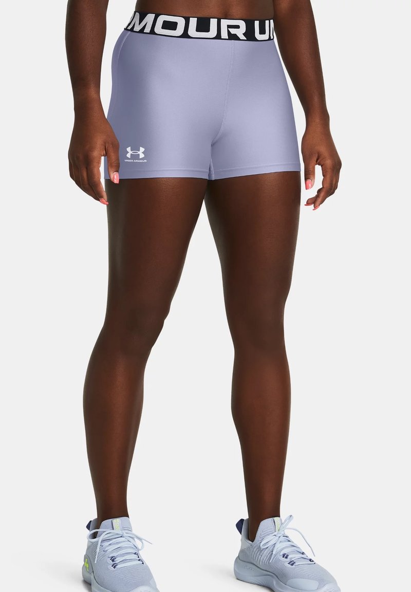 Personne en forme portant un short de compression Under Armour violet clair avec une ceinture noire et des chaussures de course gris clair, debout sur un fond blanc.