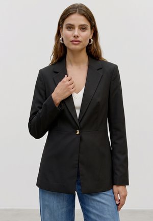 Femme aux longs cheveux bruns portant des boucles d'oreilles anneaux en argent, un blazer noir avec un bouton doré, un haut blanc et un jean bleu, debout devant un fond uni.