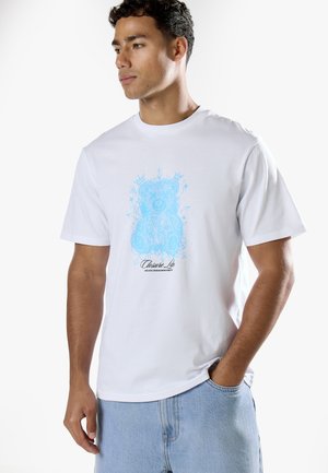 Jeune homme portant un t-shirt blanc avec un motif d'ours en peluche bleu clair et un pantalon bleu clair, debout avec une main dans la poche.