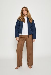 Jūras spēku zelta krāsā pūkains bomber jakas ar teksturētu virsmu, ar rāvējslēdzēju priekšpusē un sānu kabatām, apvienots ar brūnām platām biksēm un siksnām sandales.