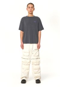 Graues Baumwoll-T-Shirt mit Rundhalsausschnitt und lila Logo. Weiße Cargo-Hose mit mehreren Taschen und Reißverschlüssen. Schwarze Schuhe.