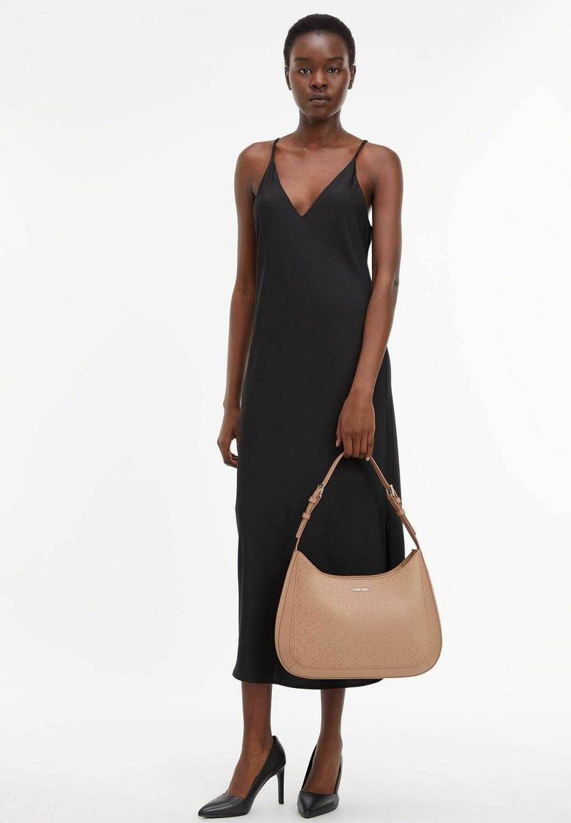 Robe noire à bretelles, sac à épaule beige avec logo en relief, chaussures noires à talons hauts, design minimaliste, texture de tissu lisse.