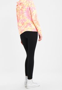 Osoba ubrana w czarne legginsy, białe sneakersy za kostkę i różowo-żółty hoodie w stylu tie-dye, stojąca na tle jednolitego jasnego tła.