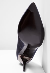 Chaussure à talon haut bleu marine avec un bout pointu et une semelle noire brillante. Le tissu texturé présente un léger éclat et une silhouette élégante.