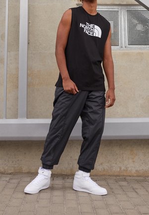 Schwarzes, ärmelloses Shirt mit weißem Logo, graue, schmal zulaufende Hose und weiße High-Top-Schuhe. Glatter Stoff, lockere Passform und elastische Bündchen an der Hose.