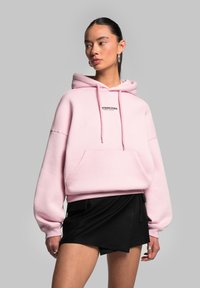Felpa oversize rosa con tasca frontale e cappuccio regolabile con cordini. Presenta un piccolo logo nero sul petto, abbinata a una gonna nera.
