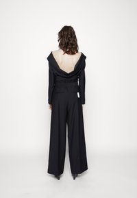 Svart jumpsuit med av-axeln-design, djup rygg, vida byxor och bälte i midjan. Slät textur och minimal varumärkesetikett synlig.