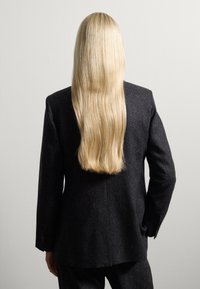 Zwarte op maat gemaakte blazer met een gestructureerde afwerking, gezien vanaf de achterkant; lange, rechte blonde haren vallen over de schouders, tegen een lichte achtergrond.