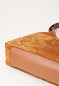 Tasche aus tanfarbenem Wildleder mit einer glatten braunen Lederunterseite. Sie zeichnet sich durch eine weiche Textur, eine strukturierte Form und verstärkte Nähte aus.