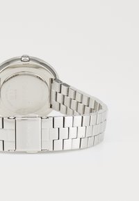Montre-bracelet en métal argenté avec un cadran rond, une lunette lisse, et un bracelet de style maillons avec une fermeture à clapet.