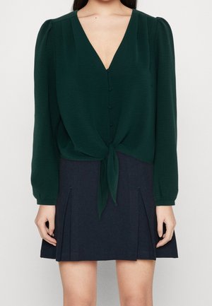 Vrouw die een donkergroene blouse met lange mouwen draagt, met knopen aan de voorkant en een detail met strik, gecombineerd met een donkere geplooide rok tegen een effen achtergrond.