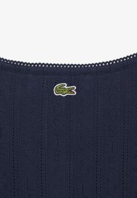 Tessuto di maglia blu navy con piccolo motivo ad occhiello e logo coccodrillo ricamato verde al centro vicino allo scollo smerlato.