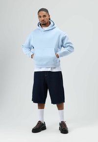 Sweat à capuche bleu clair avec une poche avant, porté par-dessus une chemise blanche. Assorti à un short en denim foncé et des baskets noires avec des lacets roses.
