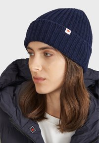 Námořnická pletená čepice s ribbovou strukturou a malou nášivkou s červeným javorovým listem. Čepice je nošena s tmavým kabátem.