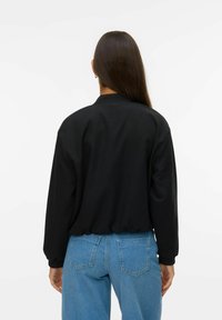 Blouson noir avec un col et des poignets côtelés, à la coupe décontractée. Porté avec un jean en denim bleu clair. Texture de tissu lisse.