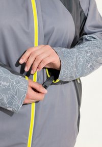 Veste grise avec une fermeture éclair jaune, des accents noirs et des manches à motifs floraux. Gros plan sur des mains qui attachent le poignet.