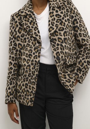 Persoon draagt een beige en zwart colbert met luipaardprint, over een wit shirt en zwarte broek, staand met één hand in de zak.