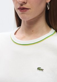 Λευκό βαμβακερό φούτερ με στρογγυλή λαιμόκοψη, διακοσμημένο με πράσινη και κίτρινη τρέσα και μικρό κεντητό λογότυπο κροκόδειλου στο στήθος.