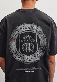Πλάτη ενός ατόμου που φοράει μαύρο T-shirt με μεγάλο ξεθωριασμένο κυκλικό λογότυπο "Pegador Football Team" σε γκρι και μικρές κόκκινες και κίτρινες λεπτομέρειες.