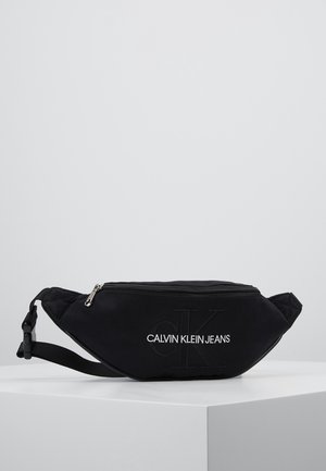 Sort Calvin Klein Jeans bæltetaske med lynlås og justerbar rem, vist på en hvid overflade mod en grå baggrund.