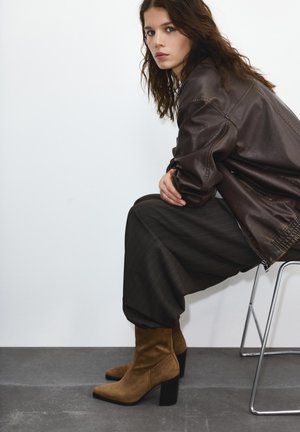 Jeune femme aux cheveux bouclés bruns portant une veste en cuir foncée, un pantalon ample et des bottes en daim marron à talons, assise sur un tabouret en métal.