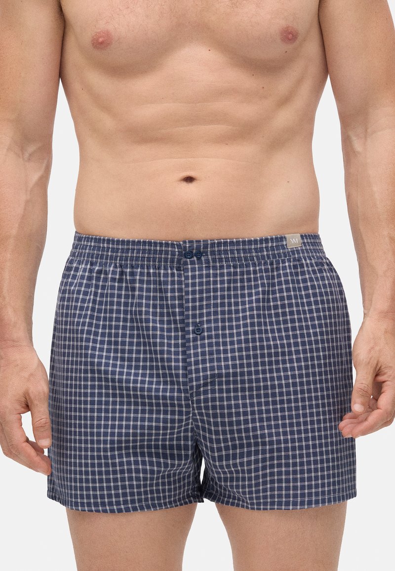 Hombre sin camiseta vestido con pantalones cortos a cuadros azules y blancos con bragueta abotonada, de pie frente a un fondo blanco liso.