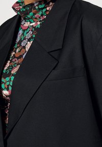 Svart kavaj med revär och en enkel bröstficka, bärs över en blommig polotröja med olika färger och mönster.