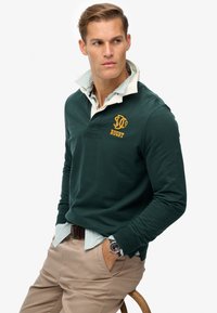 Mørkegrøn rugbytrøje med hvid krave, prydet med gul broderi. Stribede manchetter synlige. Modellen bærer beige bukser og et ur.
