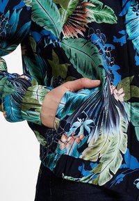 Veste bomber florale avec de grandes feuilles vertes dans des tons bleus et roses, sur un fond bleu marine sombre et des poignets élastiques.
