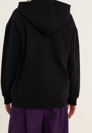 Osoba ubrana w czarną bluzę z kapturem i luźne fioletowe spodnie, widziana od tyłu na jasnym, jednolitym tle.