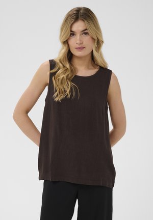 Femeie blondă cu păr ondulat, purtând un top maro închis fără mâneci și pantaloni negri, stând cu mâinile la spate.