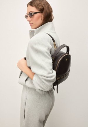 Donna con outfit grigio chiaro che indossa uno zaino in pelle marrone scuro e occhiali da sole, in piedi di profilo su uno sfondo semplice.