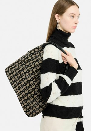 Black and beige tote bag with a repeating logo pattern, featuring a structured shape and shiny black handles. Worn over a striped sweater.