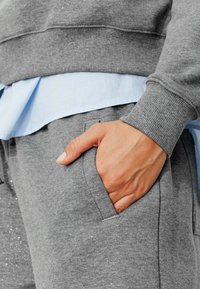 Sweatshirt gris avec des paillettes discrètes, manches côtelées, superposé à une chemise bleu clair, associé à un pantalon de survêtement gris avec poches latérales.