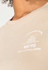 Béžové bavlněné tričko s kulatým výstřihem a potištěným bílým textem s detaily značky na levé části hrudi. Hladká textura.