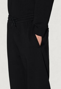 Hombre con pantalones negros de corte relajado y cintura con cordón, mano en el bolsillo lateral, vestido con una camiseta de manga larga negra a juego.