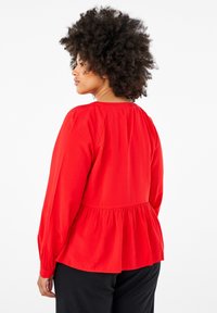 Blusa rossa a maniche lunghe con dettaglio arricciato sul retro, vita peplo e tessuto testurizzato. Presenta un colletto rotondo e polsini aderenti.