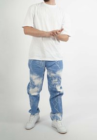 Camiseta blanca de cuello redondo, vaqueros azul claro con estampados de nubes y rayos, combinados con zapatillas blancas, de pie sobre un fondo liso.