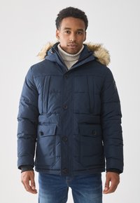 Chaqueta acolchada azul marino con capucha forrada de piel, que cuenta con cuatro bolsillos frontales, cierre de botones y textura acolchada. Llevada sobre un suéter con canalé.
