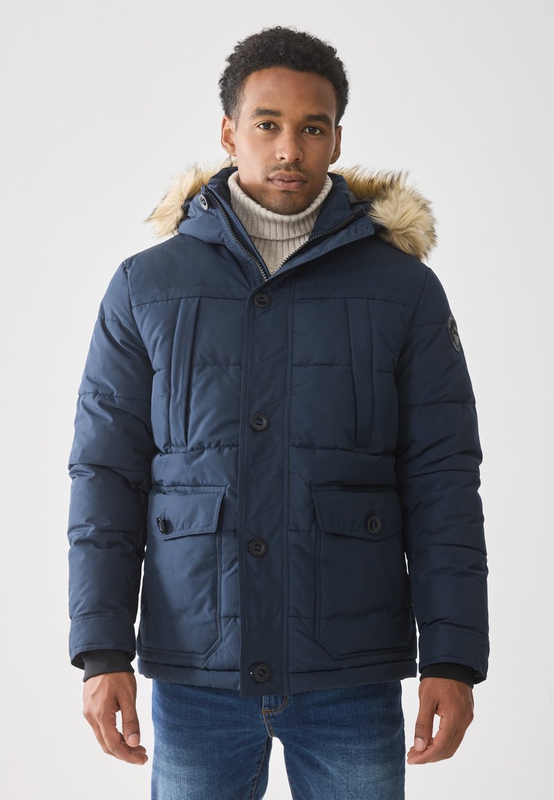 Chaqueta acolchada azul marino con capucha forrada de piel, que cuenta con cuatro bolsillos frontales, cierre de botones y textura acolchada. Llevada sobre un suéter con canalé.