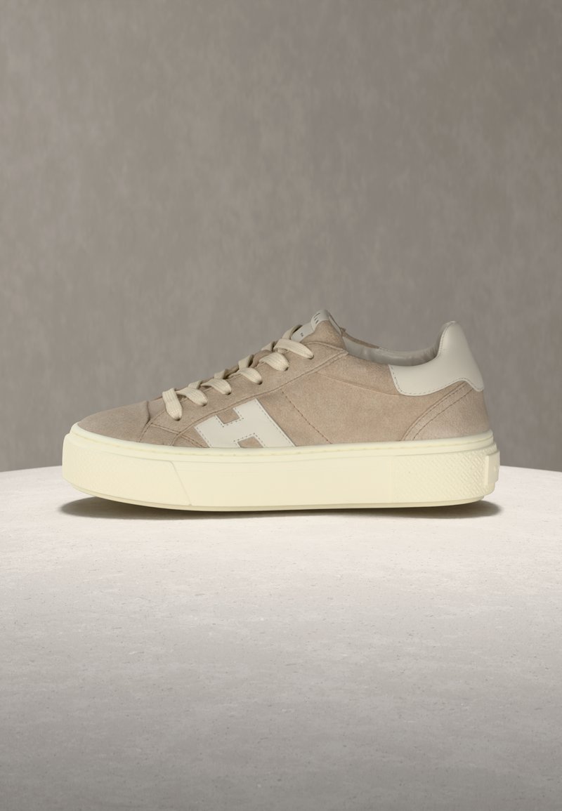 Baskets en daim beige avec lacets crème, semelle blanche épaisse et logo "H" blanc sur le côté, placées sur une surface claire contre un fond gris.