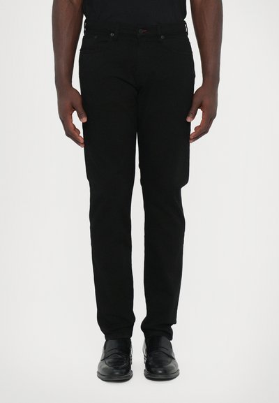 Black denim jeans with a slim fit, features five pockets, belt loops, and a button closure. Worn with black dress shoes, hands at sides.