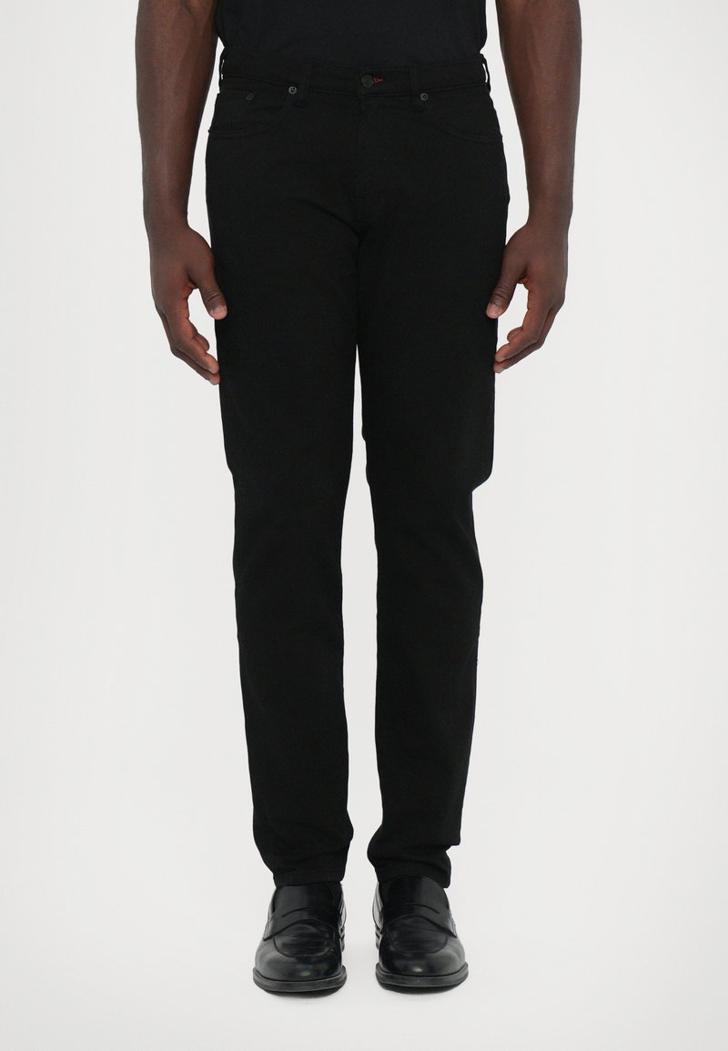 Fekete farmernadrág, slim fit szabással, öt zsebbel, övhurkokkal és gombos zárással. Fekete elegáns cipővel viselve, kezek a test mellett.