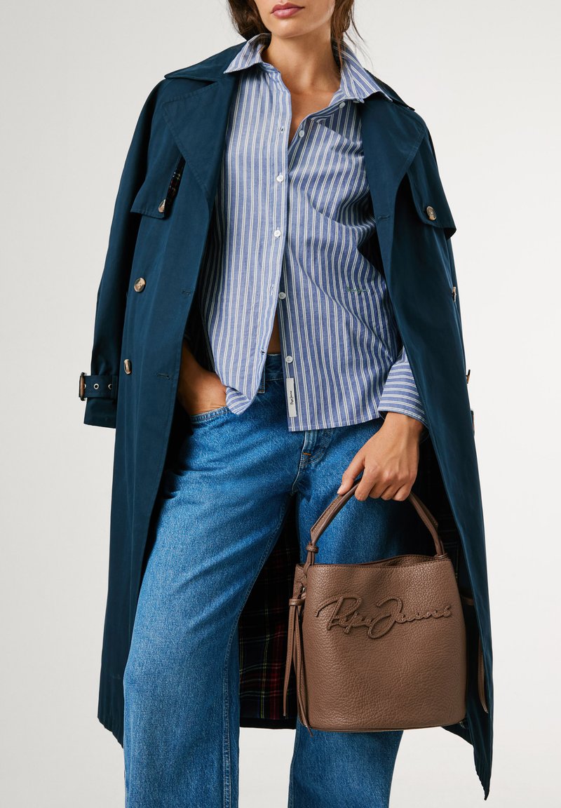 Marineblauwe trenchcoat over een blauw-wit gestreept shirt, gecombineerd met hoog opgesneden denim en een bruine gestructureerde leren handtas.