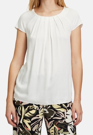 Frau trägt eine weiße Kurzarmbluse mit Falten am Ausschnitt und schwarze Hose mit Blumenmuster.